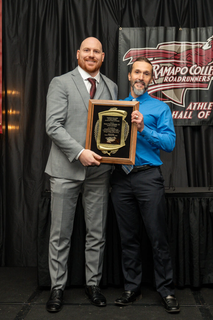 Craig Van Leeuwen and his coach Branko Miric stand on stage, smiling and holding a large plaque. One wears a grey suit and the other a blue shirt. They are on a stage with a black curtain and a Ramapo College Roadrunners banner behind them.