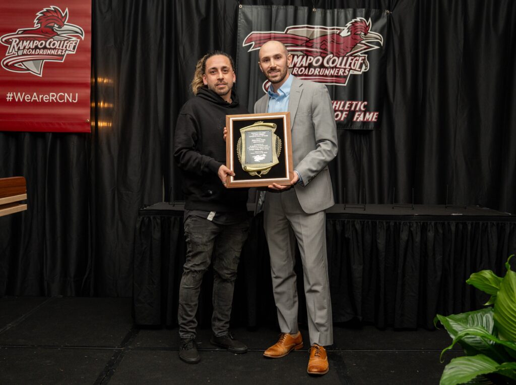 Jonathan Yazo and his former teammate Rob Stantaniello stand on a stage at a Ramapo College event. One holds a plaque, while the other stands beside him. Both face the camera, and Ramapo College banners hang behind them. A plant is visible in the lower right corner.