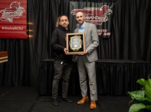 Jonathan Yazo and his former teammate Rob Stantaniello stand on a stage at a Ramapo College event. One holds a plaque, while the other stands beside him. Both face the camera, and Ramapo College banners hang behind them. A plant is visible in the lower right corner.