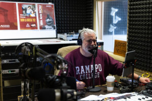 A man in a Ramapo College sweatshirt speaks into a microphone in a radio studio, surrounded by equipment. Posters and a WRPR 90.3 FM sign are visible in the background. A coffee cup sits on the desk.