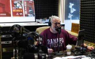 A man in a Ramapo College sweatshirt speaks into a microphone in a radio studio, surrounded by equipment. Posters and a WRPR 90.3 FM sign are visible in the background. A coffee cup sits on the desk.