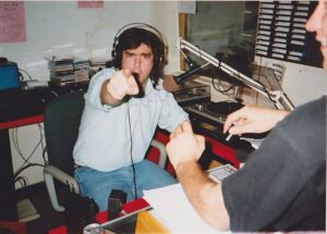 A person wearing headphones and a light shirt sits at a radio station desk, pointing toward the camera. 
