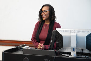 A woman stands behind a desk and smiles as she talks. A white board is behind her.