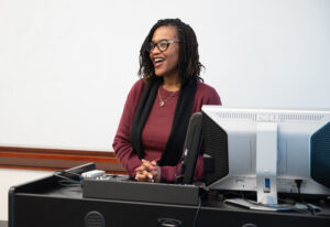 A woman stands behind a desk and smiles as she talks. A white board is behind her.