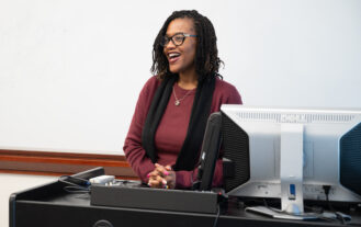 A woman stands behind a desk and smiles as she talks. A white board is behind her.