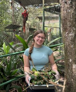 A woman with glasses and a braid, wearing a light blue shirt and gloves, holds a tray of fruit in front of a tree and outdoor animal enclosure with a red parrot perched inside. Dense greenery surrounds the scene.