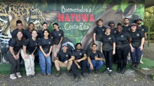 A group of people in black shirts pose and smile in front of a mural with a jaguar and the text Bienvenidos a Natuwa Costa Rica. The group stands on gravel and turf outside.