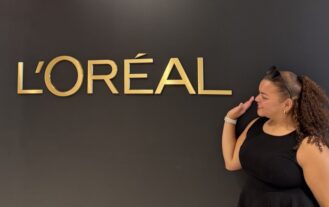 A woman stands in front of a black wall with large gold letters spelling L'OREAL