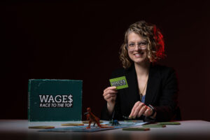 A woman smiles and holds a game card. She is seated next to a box that states: WAGES.