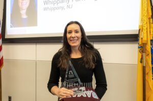 A woman with long dark hair, wearing a black top, smiles while holding a clear triangular award. Behind her, a presentation slide and yellow flags are visible.