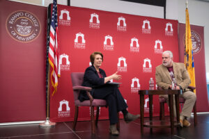 Two people sit and talk on stage at Ramapo College of New Jersey, with red banners and flags behind them. A woman holds a pen and paper, while a man wearing a blazer listens.