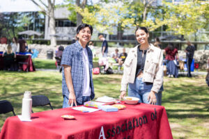 Two college students stand outside behind a table with a red tablecloth.