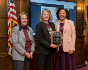 Three women stand side by side. One is holding an award.