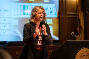 A woman stands with a microphone in her hand and speaks. She is in front of a screen.