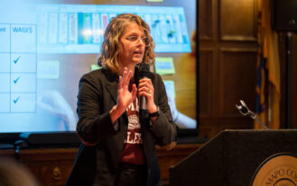 A woman stands with a microphone in her hand and speaks. She is in front of a screen.