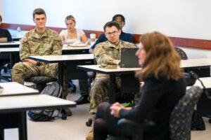 Students sit at tables in a classroom. Two are wearing army fatigues. All are looking at guest speaker Mary Legere in the foreground.