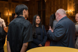 A group of people in formal attire stand and converse in an elegant, wood-paneled room with warm lighting and a yellow tablecloth in the foreground.
