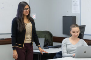 Two women in a classroom: one stands and smiles near a desk, while the other sits, looking at her laptop. There are desks, a whiteboard, and office equipment in the background.