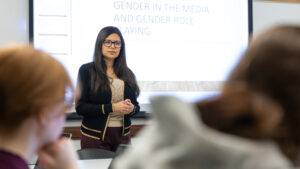 A woman stands at the front of a classroom, speaking to students. Behind her, a projected presentation reads Gender in the Media and Gender Role-Playing. Two students are visible in the foreground.