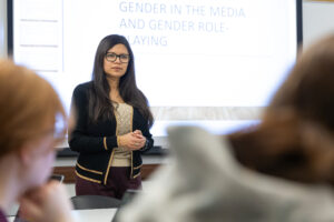 A woman stands at the front of a classroom, speaking to students. Behind her, a projected presentation reads Gender in the Media and Gender Role-Playing. Two students are visible in the foreground.