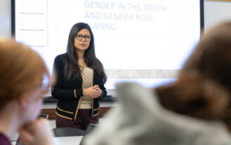 A woman stands at the front of a classroom, speaking to students. Behind her, a projected presentation reads Gender in the Media and Gender Role-Playing. Two students are visible in the foreground.