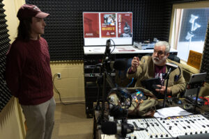Two men are in a soundproof radio studio; one stands by the door listening while the other sits at a desk with audio equipment, speaking and gesturing with a pen. Posters and a window are visible in the background.