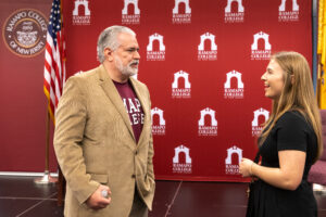 A man in a beige blazer and maroon shirt talks with a smiling woman in black, standing in front of a Ramapo College of New Jersey backdrop, with a U.S. flag and college seal visible.