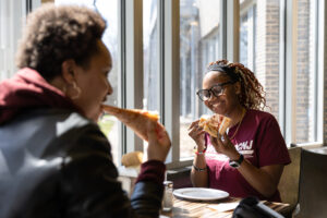 Two students sit at a table next to a window in the Birch Tree Inn dining hall on the RCNJ campus eating pizza.