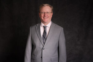 A man wearing glasses, a light gray suit, a light pink shirt, and a striped tie stands smiling in front of a dark, textured background.