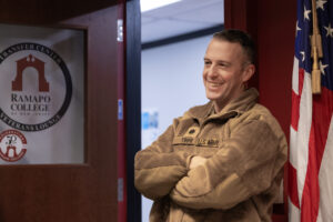 A person wearing army fatigues stands outside the Ramapo College Veterans lounge with arms folded next to an American flag. The door shows the Ramapo College logo of the Arch with the words "transfer students and veterans lounge in a circular position.