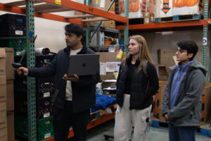 Three people stand in a warehouse surrounded by boxes and crates. One person holds a laptop and scans boxes with a barcode reader while the other two watch attentively. Shelves with various items are in the background.