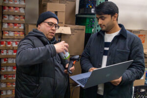 Two people stand in a storage room with shelves of canned goods; one scans a can with a handheld device while the other holds a laptop and observes.