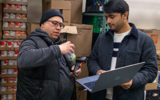 Two people stand in a storage room with shelves of canned goods; one scans a can with a handheld device while the other holds a laptop and observes.