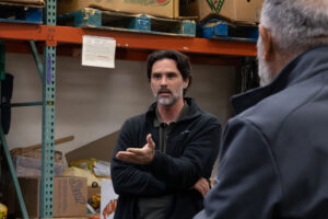 A man with dark hair and a beard gestures while speaking in a warehouse, with shelves of boxes and food items behind him. Another person, seen from behind, listens in the foreground.