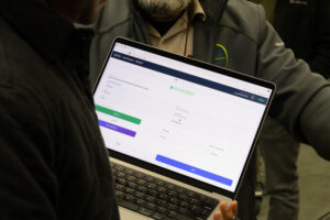 A person holds a laptop displaying a web application form, while another person stands nearby. The screen shows fields and buttons in white, green, and blue. Both people are wearing jackets.