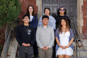 Six students are standing on stairs outside of Birch Mansion on campus. There are three in each of two rows.