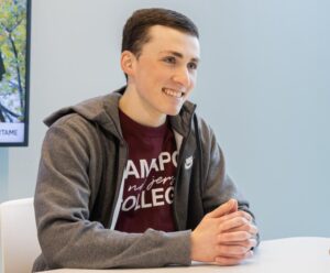 A young man wearing a Ramapo College shirt sits and smiles.