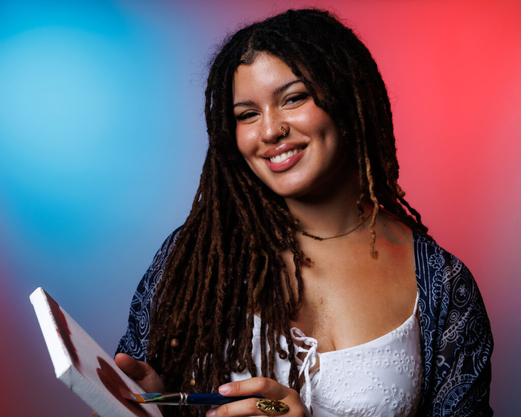 Chantalle Reyes is smiling and holding a paint canvas and paint brush. The canvas shows a painted heart. The background is a blue and red gradient backdrop,