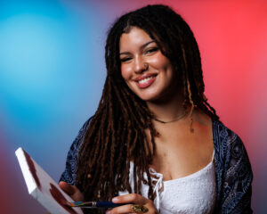 Chantalle Reyes is smiling and holding a paint canvas and paint brush. The canvas shows a painted heart. The background is a blue and red gradient backdrop,