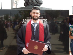 A man in  black graduation gown and maroon stole holds a diploma.