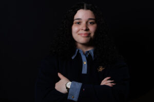Emally Quezada is standing with arms crossed in front of a dark background, smiling.