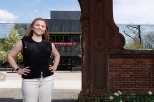 A young woman stands outside under a historic arch with a building in the backgound.