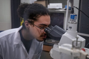 A young man wearing a lab coat and glasses looks through a microscope.