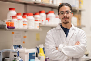 A young man in a white lab coat stands with his arms crossed in a lab.