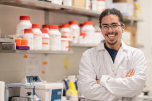 A man with a white lab coat on stands in a lab with his arms crossed. He is smiling.