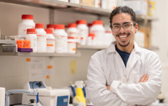 A man with a white lab coat on stands in a lab with his arms crossed. He is smiling.