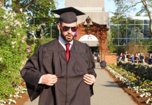 A man in a black cap and gown walks outside. A large arch is behind him.