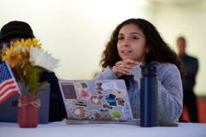 A young woman sits in front of a laptop with stickers on it. Her hands are folded and she is looking up.