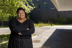 A woman stands outside under a building's overhang with her arms crossed and smiling.