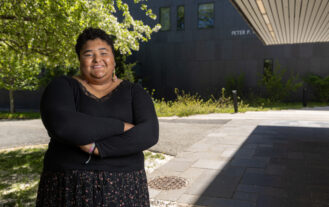A woman stands outside under a building's overhang with her arms crossed and smiling.